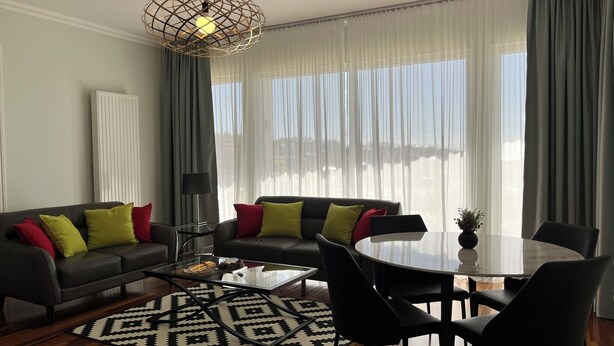 Un salon moderne avec un canapé, une table basse et des fauteuils, un tapis à motifs et une grande fenêtre avec des rideaux.