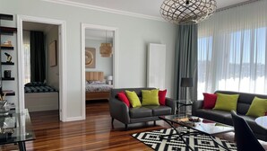 Un salon moderne avec un canapé en cuir noir, une table basse en verre et un tapis à motifs. On aperçoit, au fond, un lit avec une tête de lit, ainsi qu’un coin repas avec une table en verre.