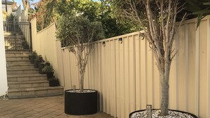 Une terrasse dallée avec deux grands bacs à fleurs noirs, une clôture beige et un escalier menant à un niveau supérieur.