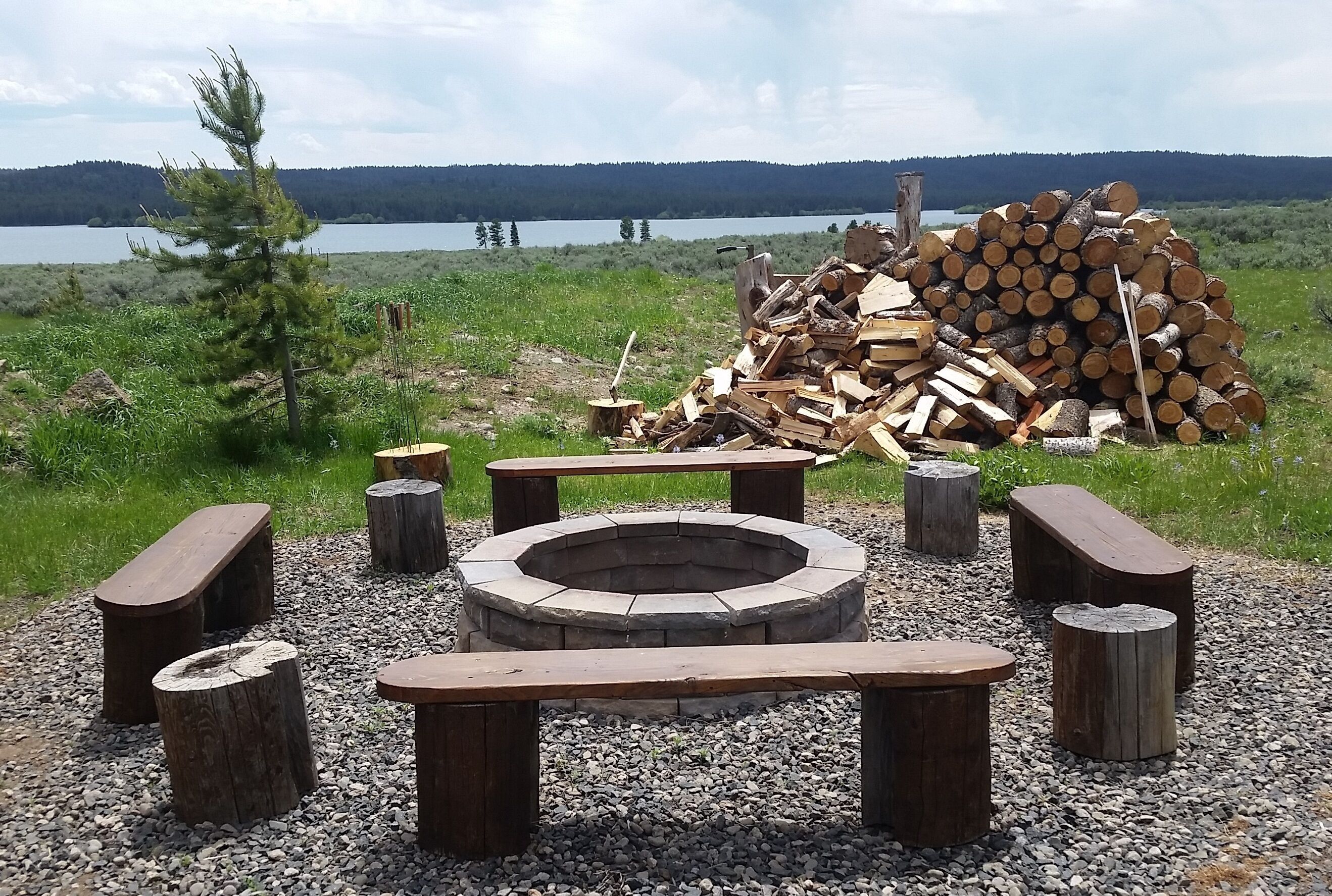 A fire pit surrounded by wooden benches and a pile of logs, set against a backdrop of a lake and forest.