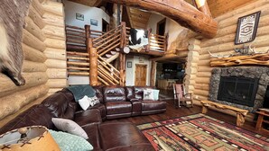 Ein Blockhaus-Interieur mit Ledersofa, einem gemusterten Teppich, einem Kamin und einer Treppe.