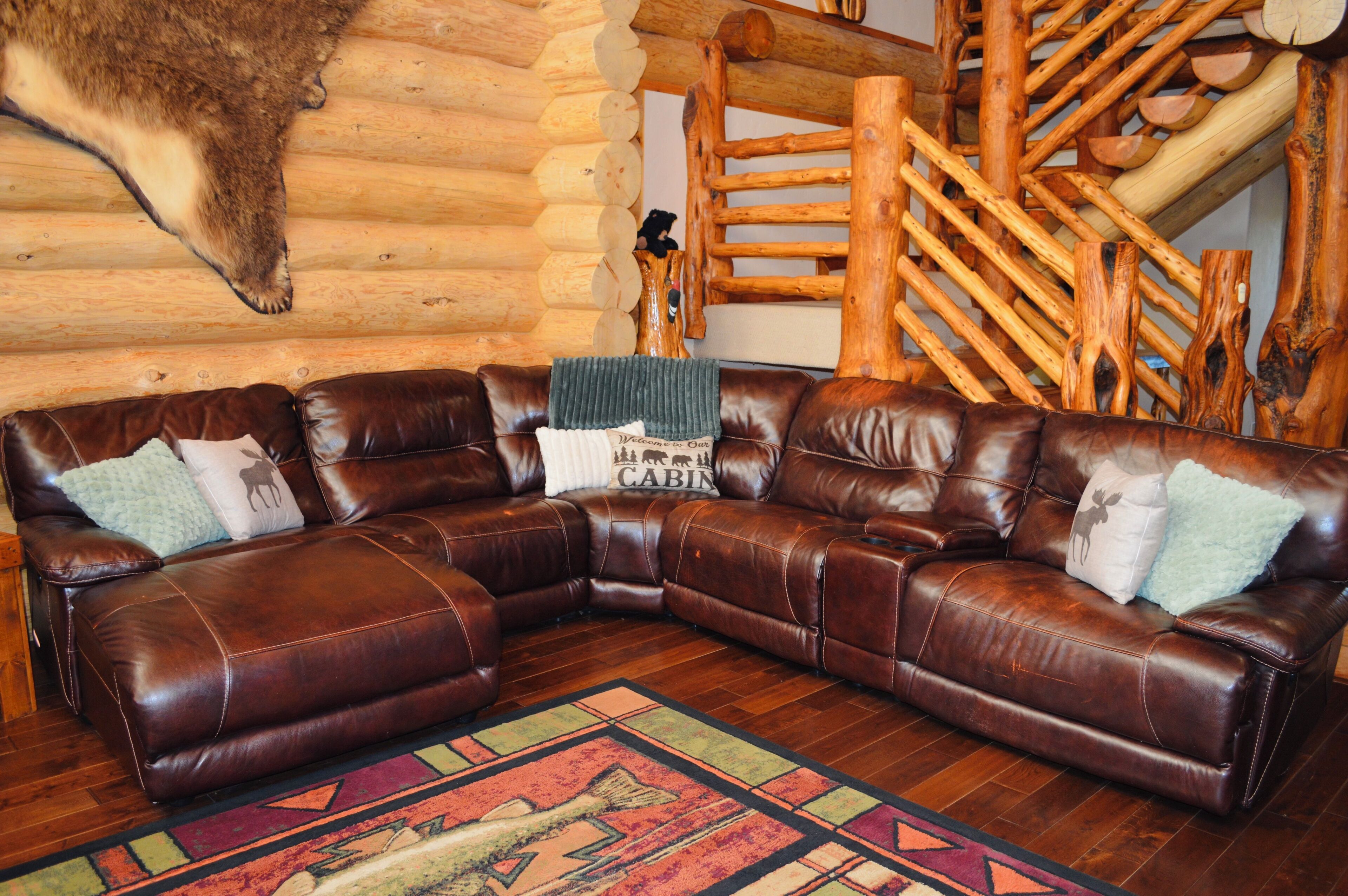 A brown leather sectional sofa with pillows, a colorful rug, and a wooden log wall with a mounted animal head.