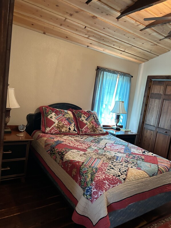 A bedroom with a wooden ceiling, a bed with a colorful quilt, a nightstand with a lamp, and a window with curtains.