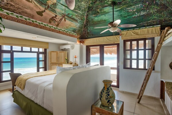 Un dormitorio con una cama grande, un mural en el techo, un ventilador de techo y vista a la playa.