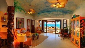 Een ruime woonkamer met een groot raam dat uitzicht biedt op de oceaan, een plafondventilator en een tropische muurschildering.