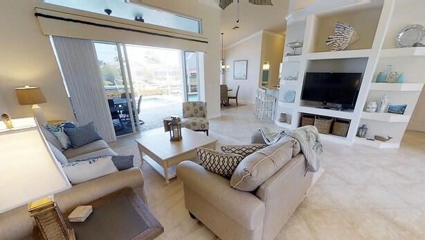 A spacious living room with a sofa, coffee table, and TV stand. There is a dining area with chairs and a table, and a large window providing a view of the outdoors. - Sunny Pool & Spa inside Lanai! Dock and Water access! 3bd 2bath home. (Marco Island)