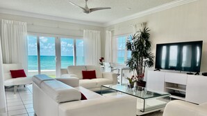 A modern living room with a large TV, white sofas, a glass coffee table, and a view of the ocean. - Regal Beach Club #614 (Seven Mile Beach)