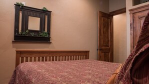A bedroom with a wooden headboard, a patterned bedspread, a mirror with greenery, and a wooden door.
