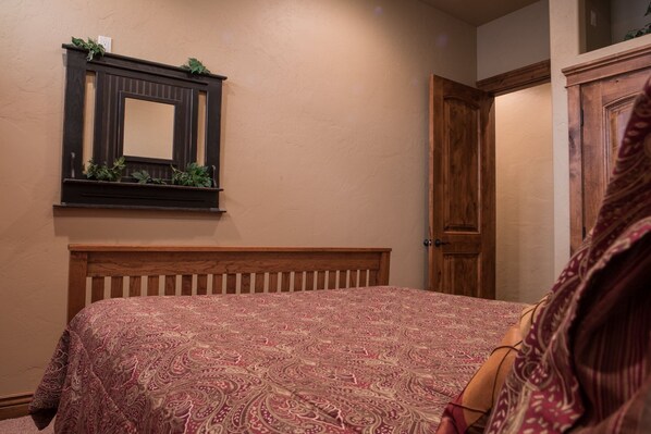A bedroom with a wooden headboard, a patterned bedspread, a mirror with greenery, and a wooden door.