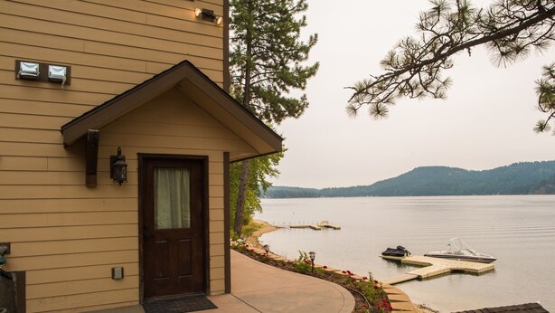 A house with a wooden exterior, a front door, and a porch leading to a lake with boats.