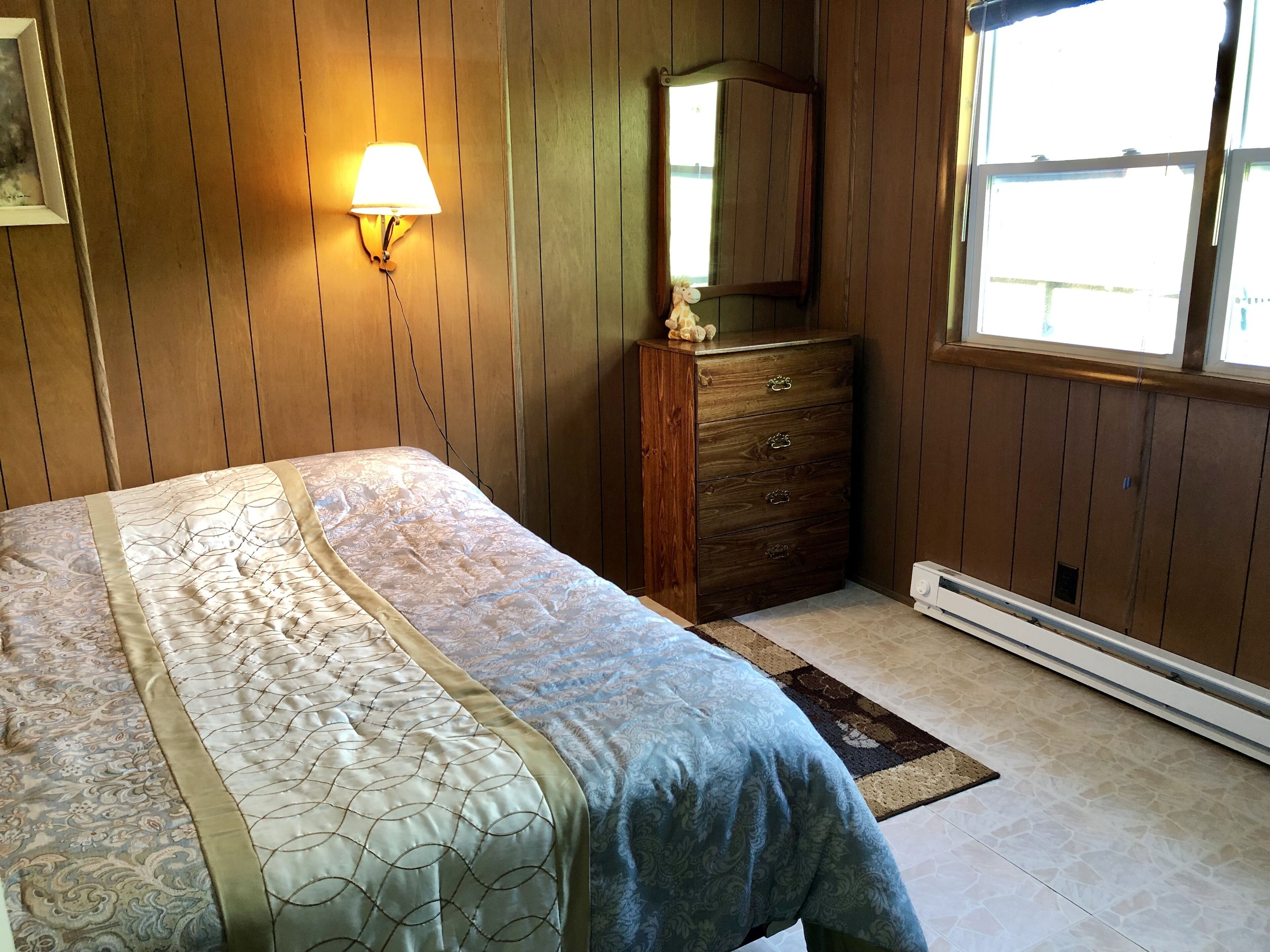 A bedroom with a bed, a dresser, a wall-mounted lamp, a mirror, and a window.