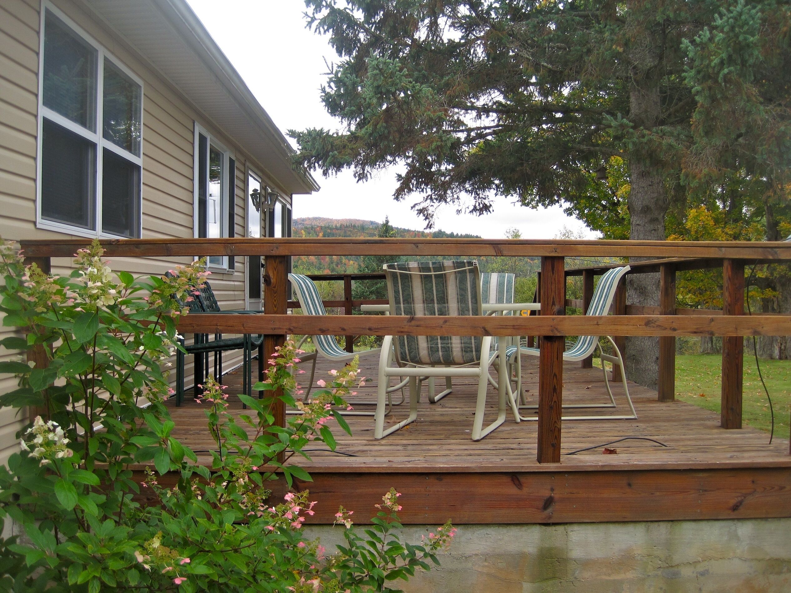 A wooden deck with outdoor furniture, including chairs and a table, overlooking a house and natural landscape.