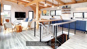 A spacious room with a high ceiling, wooden beams, a dining area, a kitchen, and a TV mounted on the wall. - Windows on the Sea ★ Modern Pool & Beach Retreat ★ 100 ft to Beach ★ 2 Masters (Ocean Beach)