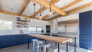 A modern kitchen with a wooden ceiling, white cabinets, stainless steel appliances, and a central island with stools. - Windows on the Sea ★ Modern Pool & Beach Retreat ★ 100 ft to Beach ★ 2 Masters (Ocean Beach)