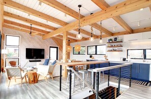 A spacious room with a high ceiling, wooden beams, a dining area, a kitchen, and a TV mounted on the wall.