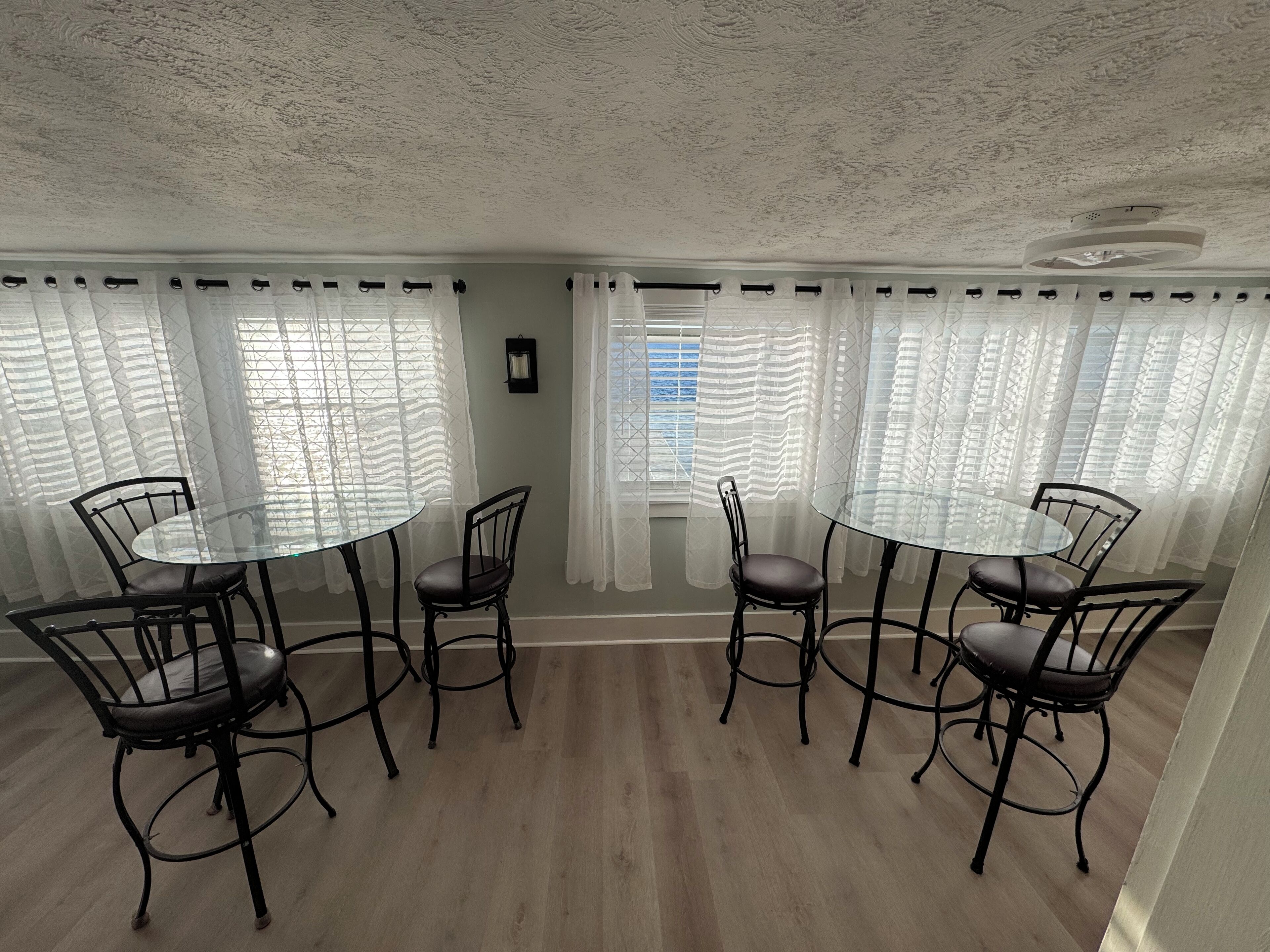 A dining area with round glass tables and black metal chairs, surrounded by white curtains and a view of the sea.