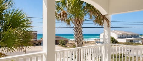 A covered balcony with a view of the ocean and beach, white railings, and a palm tree.