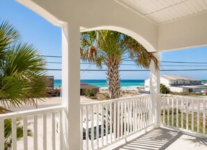 A covered balcony with a view of the ocean and beach, white railings, and a palm tree.