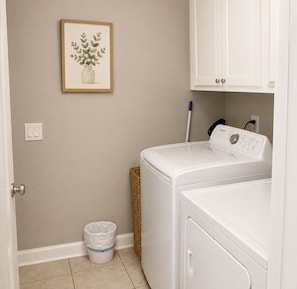 Une buanderie équipée d’un lave-linge, d’un sèche-linge, d’un panier en osier, d’une poubelle et d’un tableau encadré accroché au mur.