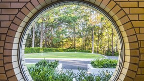 A view through a circular window with a brick frame, displaying a well-maintained lawn, a curved pathway, and a dense tree line in the background. - Fern Ridge Private Resort/ Wedding / Function Venue (Coffs Harbour)