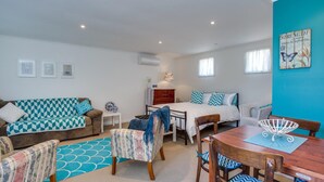 A cozy, well-lit room with a sofa, a bed, a dining table, and a chair. There are framed pictures on the wall, a small table with a lamp, and a decorative wall piece.