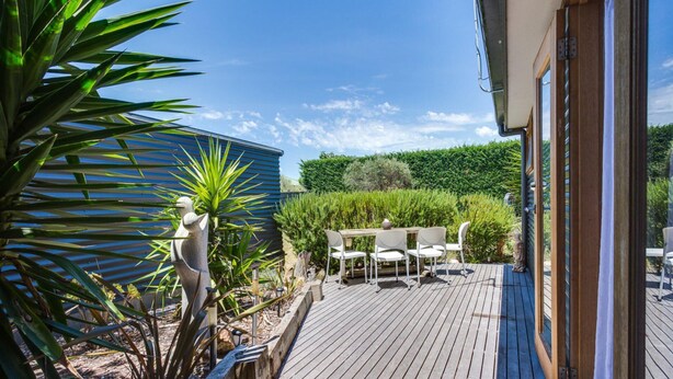 A wooden deck with a table and chairs, surrounded by greenery and a blue building.
