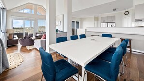 A spacious living area with a large white table and blue chairs, a kitchen in the background, and a balcony with a view. - Flinders View Beach House (4Bed 2Bath) - Flinders View Beach House - Executive Holiday Home (Augusta)