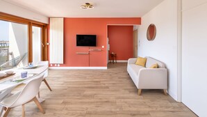 A modern living room with a white sofa, a dining table with chairs, a flat-screen TV mounted on the wall, and a large window offering a view of the city. - 🌴 Sunny 3-Bedroom Marina Apartment | Sea Views | Secure Parking | Beach Walk (Mauguio)