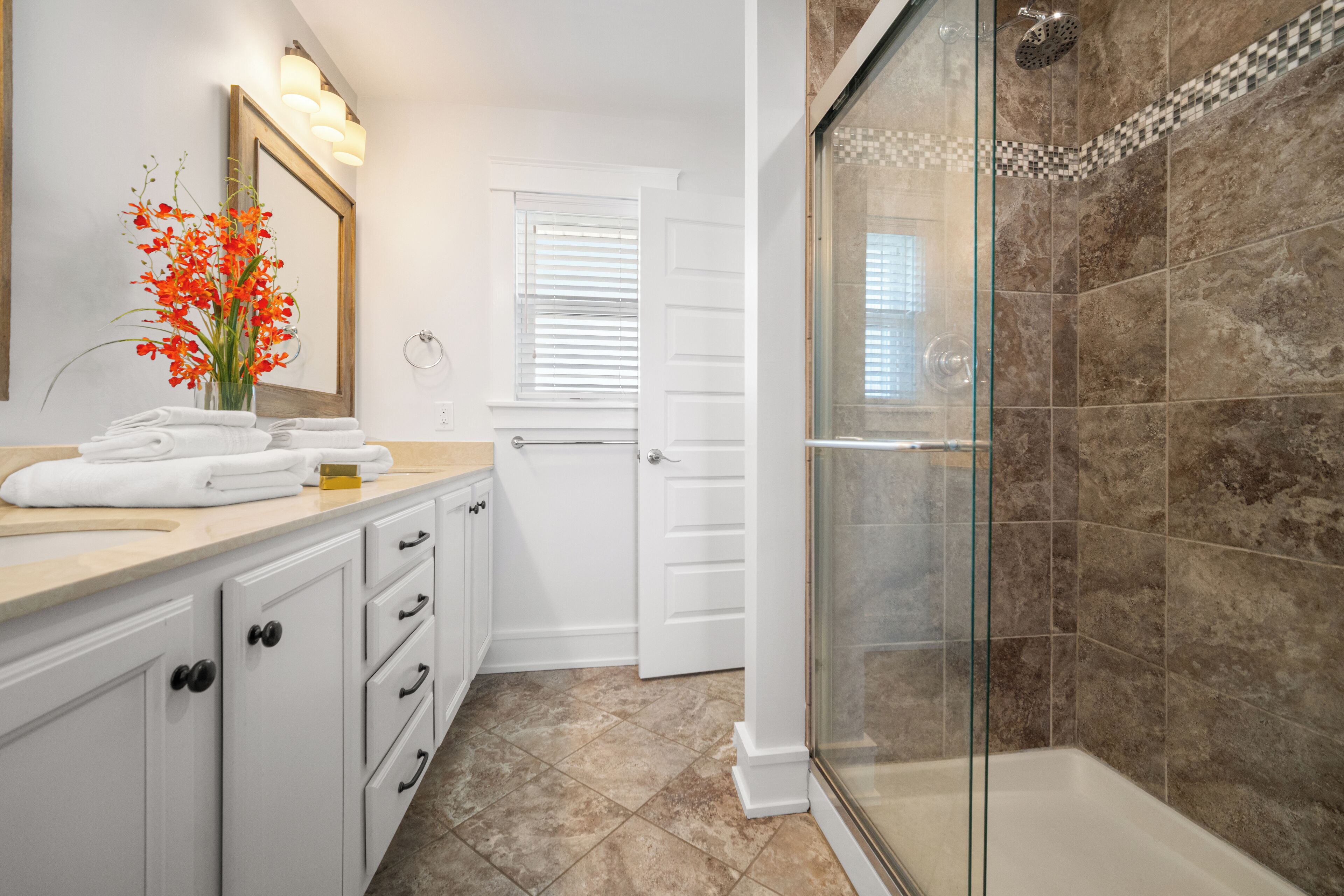 Un baño con cabina de ducha de vidrio, muebles de tocador blancos y un florero con flores rojas en la encimera.