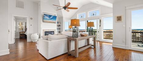 A spacious living room with hardwood flooring, a ceiling fan, and large windows offering a view of the ocean.