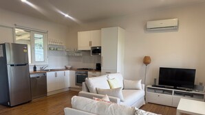 Een moderne keuken met witte kasten, een roestvrijstalen koelkast en een magnetron. In de woonruimte bevindt zich een witte bank met kussens, een flatscreen-tv en een kleine tafel.