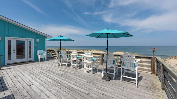 Une terrasse en bois, agrémentée de chaises Adirondack blanches et de parasols turquoise, donnant sur une plage.