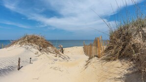 Un sentier de sable qui longe la plage et mène à l’océan, bordé de barrières en bois et parsemé de quelques plantes.