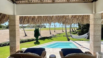 A covered outdoor area with a pool, wicker furniture, and a view of a landscaped garden.