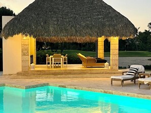 Un área junto a la piscina con una estructura de techo de paja, sillas de descanso y una piscina azul cristalina.