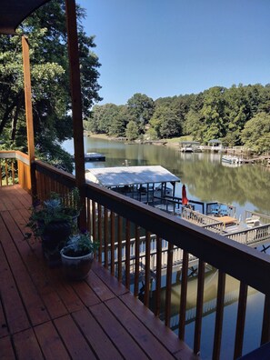 Una terraza de madera con plantas en macetas, con vista a un lago tranquilo con barcos amarrados y un pabellón.