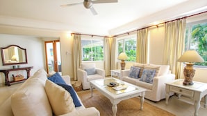 A spacious living room with a white sofa, a coffee table, and a TV. There are curtains, a ceiling fan, and a window with a view of greenery. - Palm Beach 109 Hastings Barbados, 3 Bdrm condo. Beachfront, Pool, Best location! (Hastings)