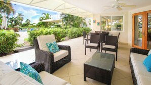 A covered outdoor seating area with wicker furniture, a glass-top table, and a view of a pool and greenery. - Palm Beach 109 Hastings Barbados, 3 Bdrm condo. Beachfront, Pool, Best location! (Hastings)