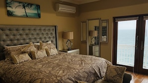 Quarto com uma cama grande, mesa de cabeceira, abajur, espelho e vista para o oceano através de uma porta.