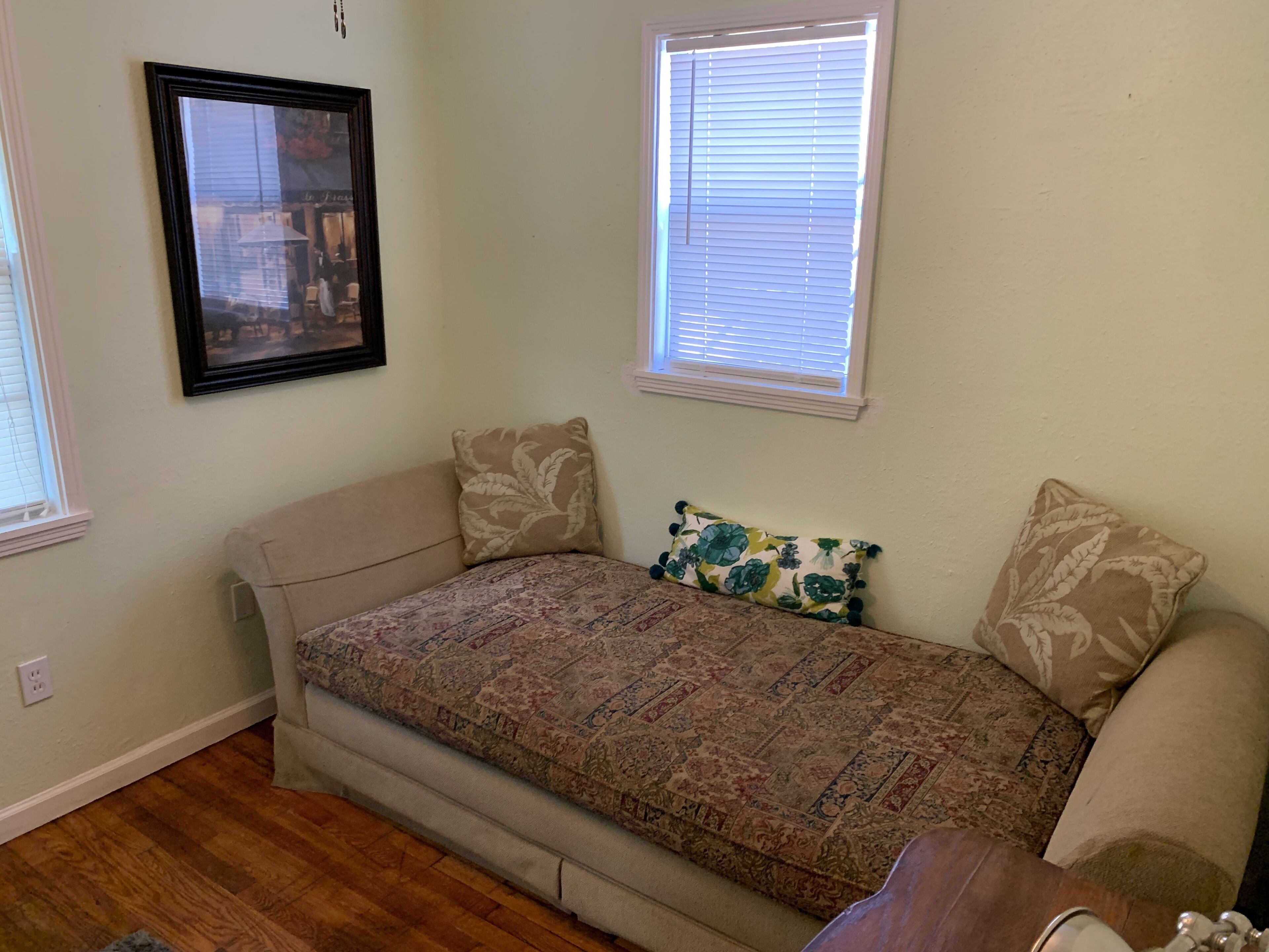 Pequeña habitación con cama individual, colcha estampada, dos almohadas, un cuadro enmarcado en la pared y una ventana con persianas.