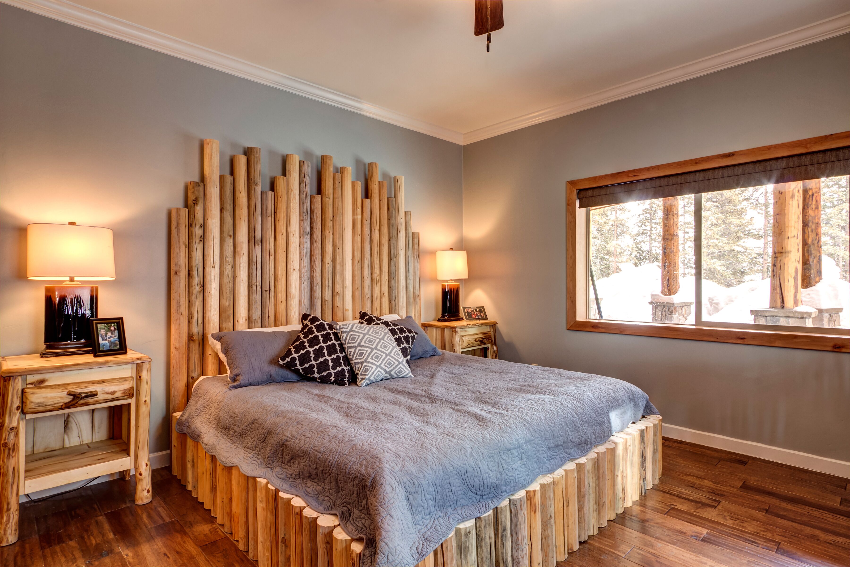A bedroom with a wooden headboard, a large bed with a blue blanket, two bedside tables with lamps, and a window with a view of trees.