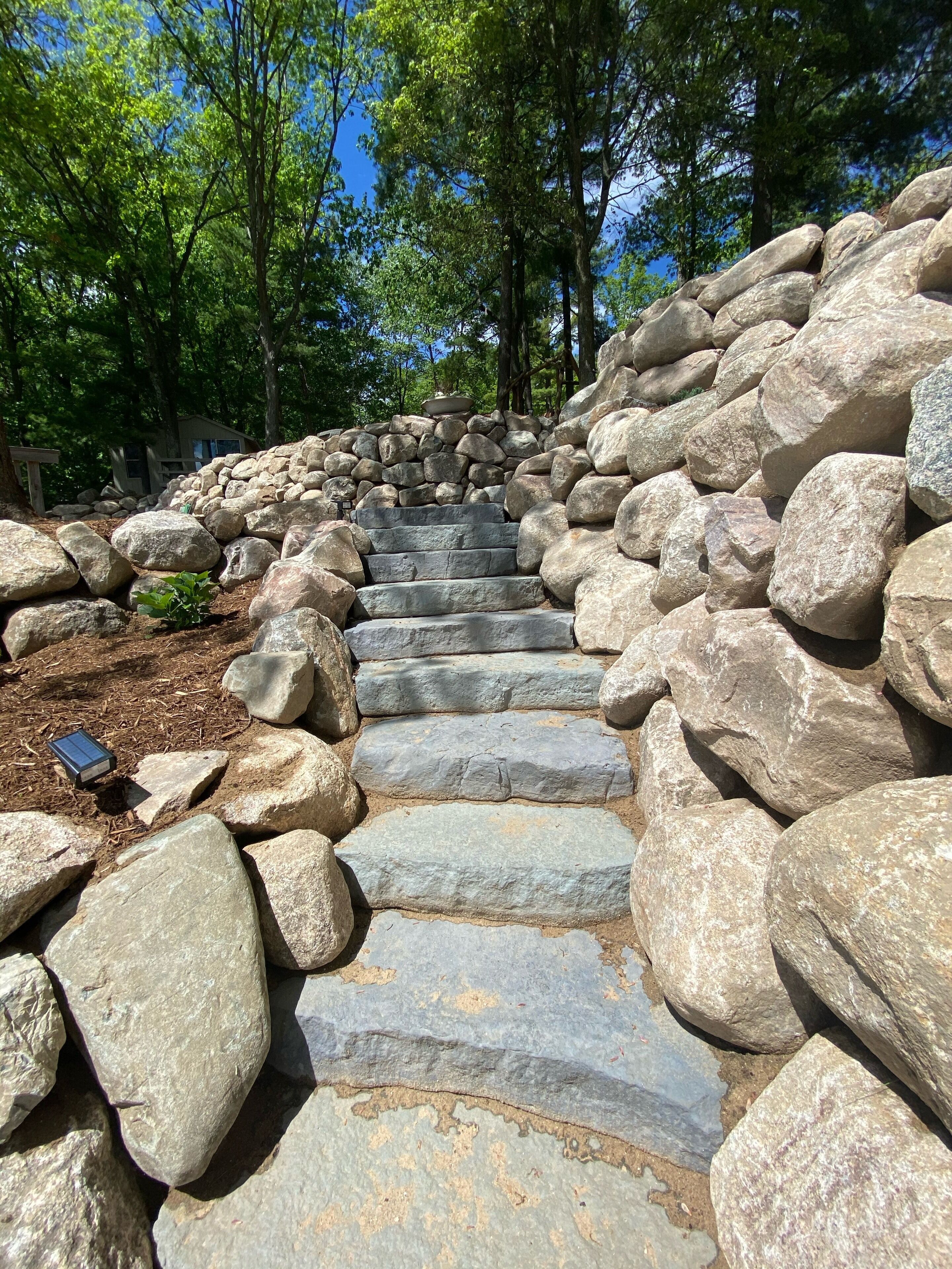Eine steinerne Treppe, umgeben von großen Felsen, die einen Hügel hinauf führt, mit Bäumen im Hintergrund.