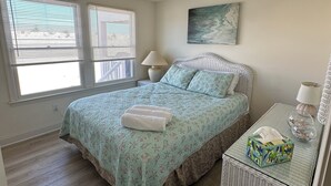 A bedroom with a bed, a bedside table, a lamp, a painting, and a window with blinds.