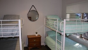 A dormitory room with bunk beds, a small wooden nightstand, a mirror, and a window with a view.