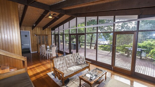 A spacious room with a large wooden ceiling, a sofa, a dining table, and a view of a deck with outdoor furniture.