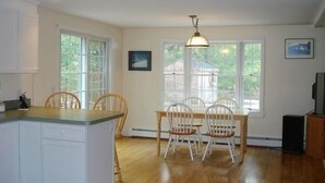Et køkken med hvide skabe, et spisebord med stole og store vinduer med udsigt til omgivelserne.