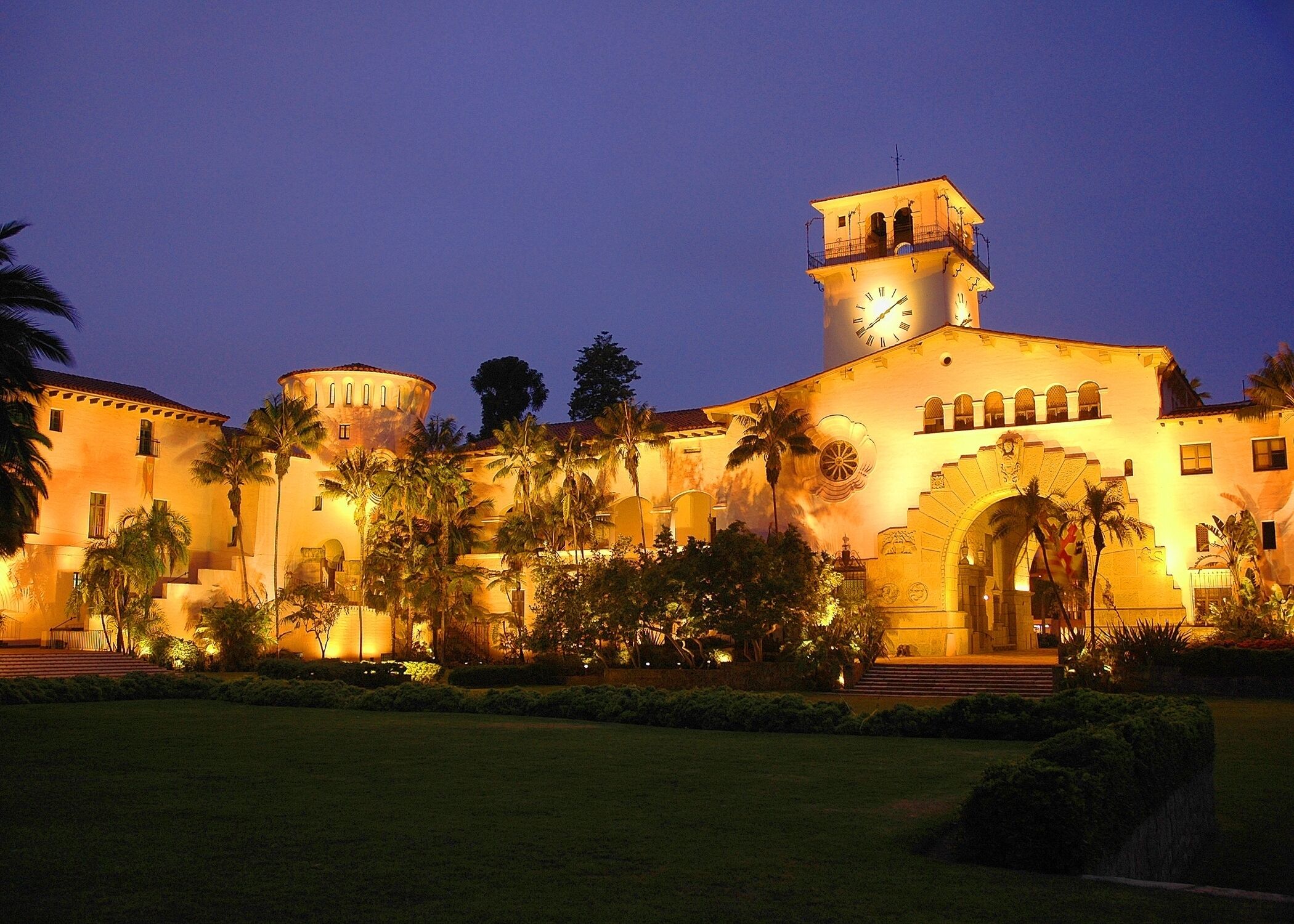 A large, historic building with a clock tower, palm trees, and a well-lit entrance.