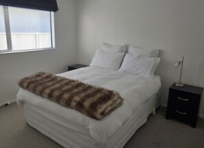 Un dormitorio con cama, ropa de cama blanca, un plaid de piel sintética, una mesita de noche, una lámpara y una ventana con persianas.