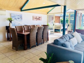 Un coin repas avec des chaises en osier et une table en bois, un canapé bleu et une vue sur l’extérieur grâce à de grandes fenêtres.