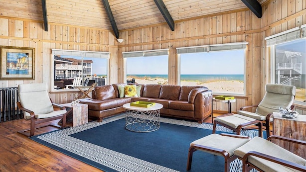Ein Holzinnenraum mit einem Ledersofa, Sesseln, einem Couchtisch und großen Fenstern mit Blick auf einen Strand.